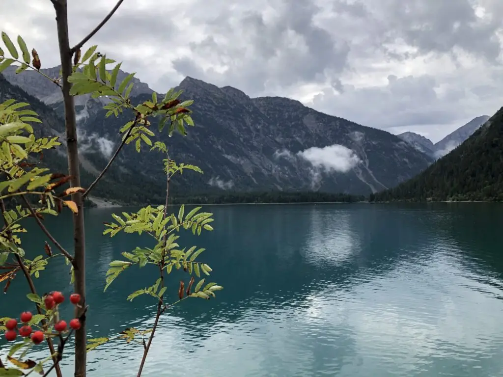 Bergsee umgeben von Bergen, bewölkte Stimmung, im Vordergrund Äste einer Ebersche mit roten Beeren.